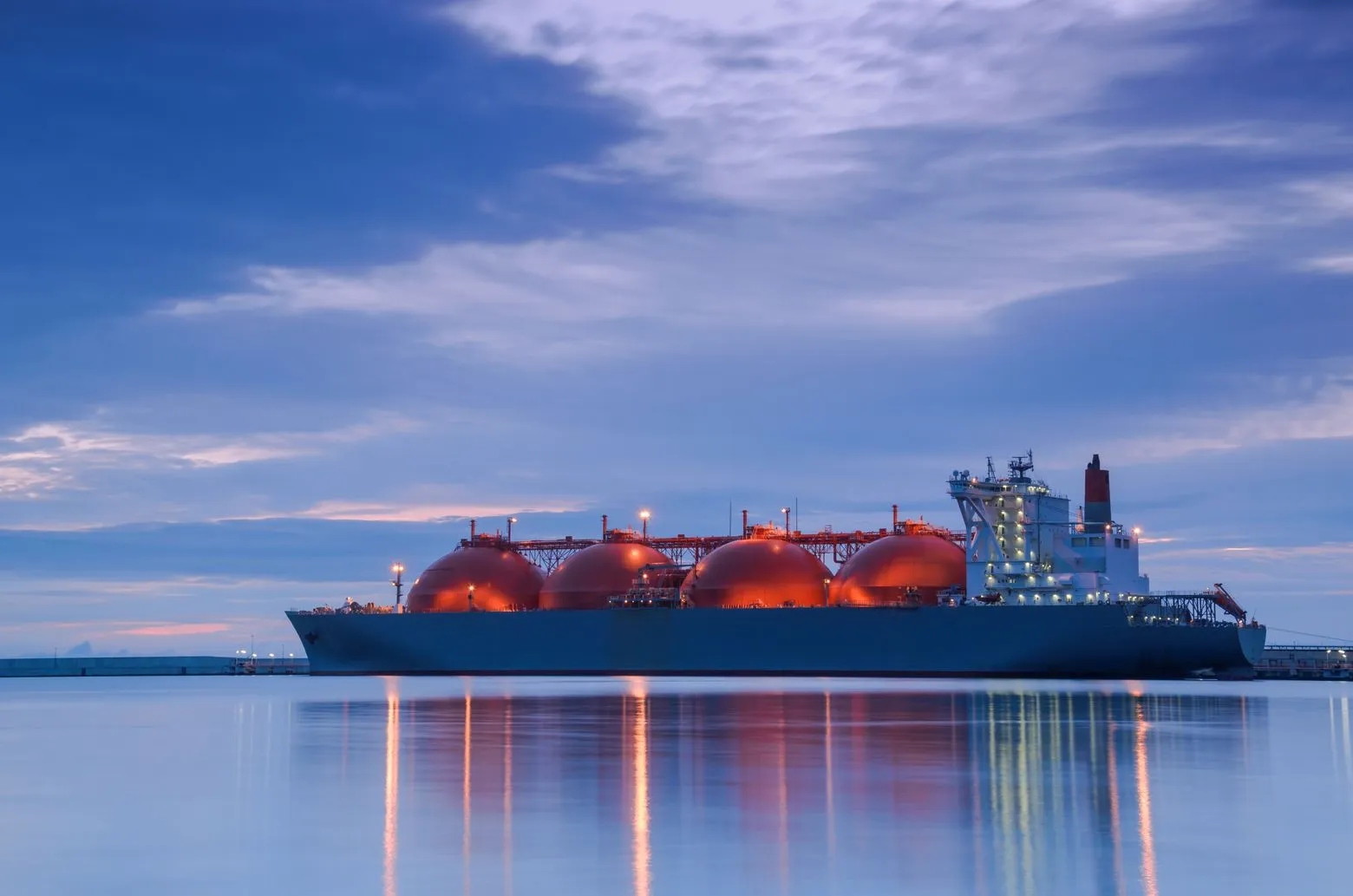 A large LNG tanker with spherical orange tanks is docked at a port during sunset, reflecting on calm water under a cloudy sky.