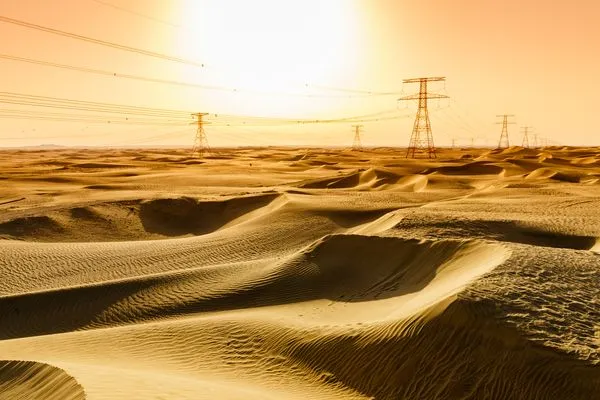 Golden desert landscape with sand dunes under a bright sun. Power lines stretch across the horizon, casting long shadows on the sand.
