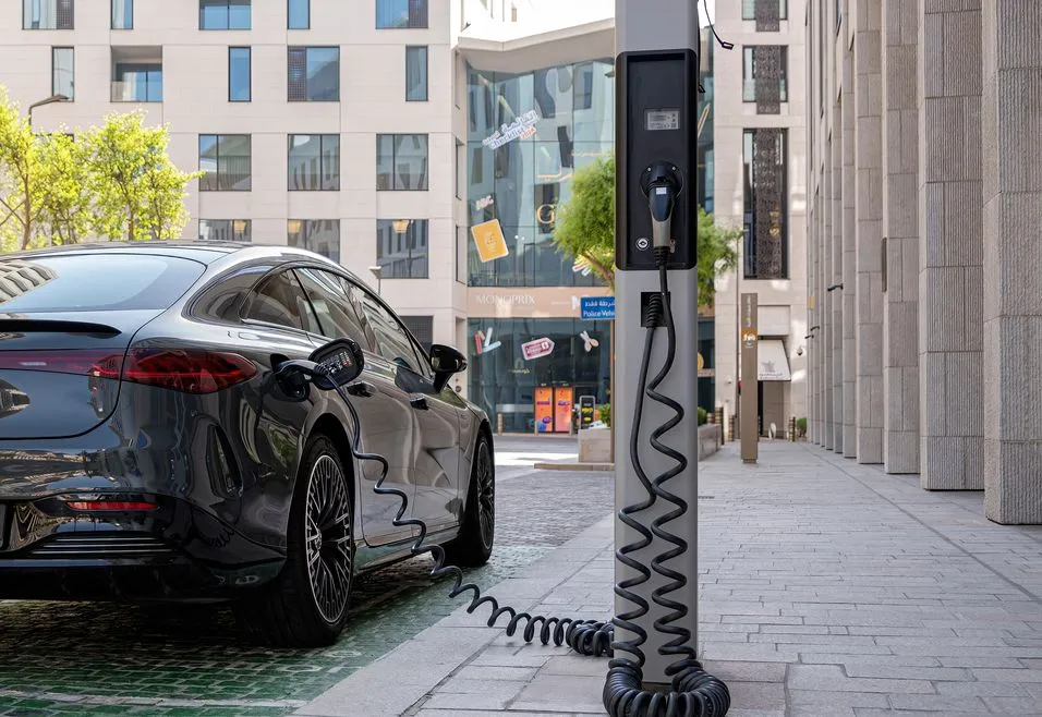 Electric car charging at a public station on a city street, surrounded by modern buildings and trees.